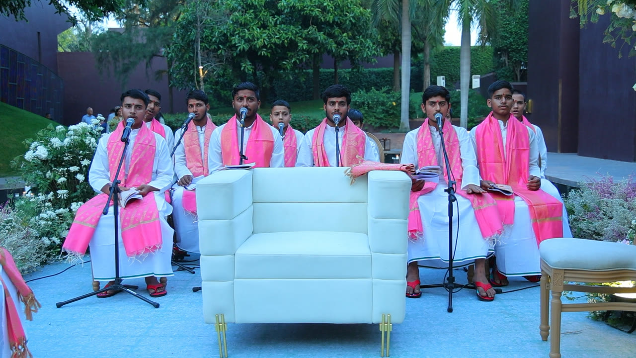 A row of young pandits in white kurtas and rose-gold angavastram chanting into microphones at a garden mandap