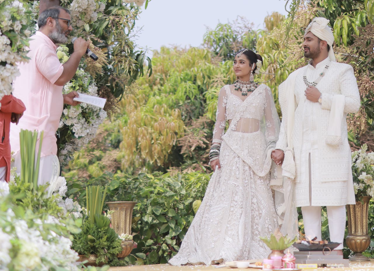 Hemant Acharya singing a mantra into a microphone while a bride and groom stand hand in hand at a garden mandap